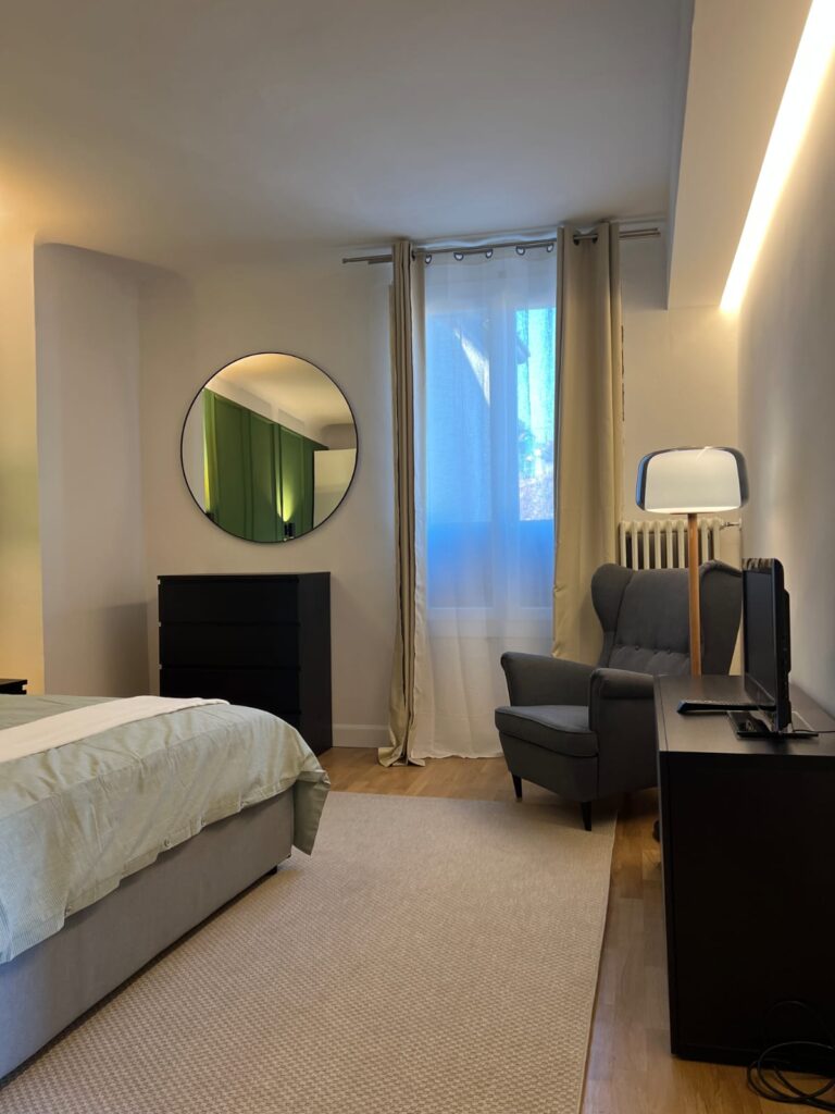 Camera da letto del bilocale a Brescia con specchio tondo, poltrona e illuminazione indiretta a soffitto